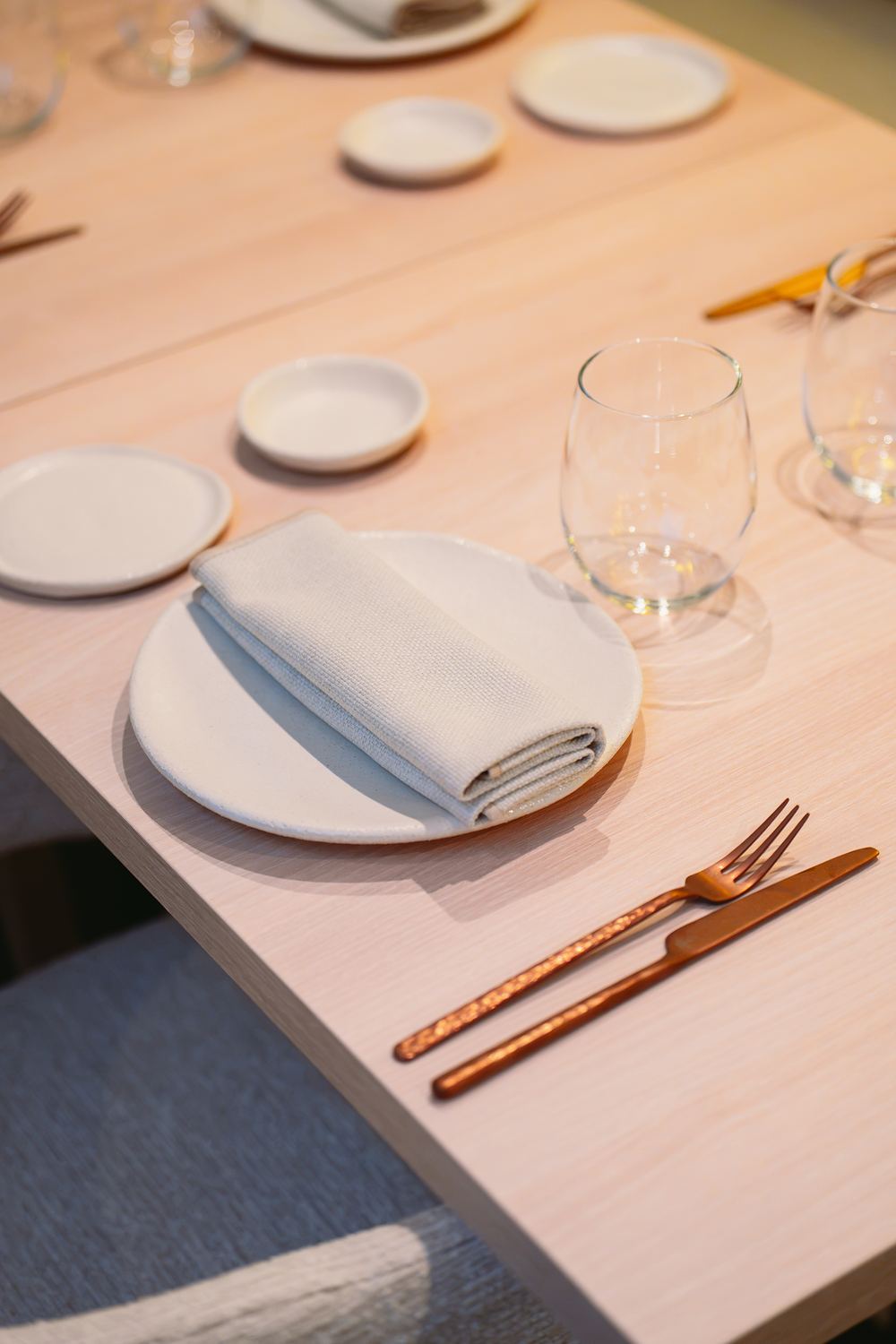 Detalle de una mesa de Nodo: plato blanco, servilleta doblada y cubiertos de cobre sobre madera clara